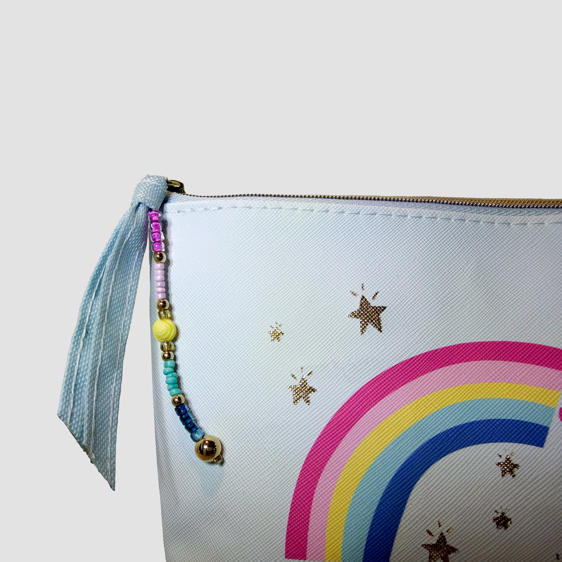 A close-up of a white zippered pouch featuring a colorful rainbow and star print, with a light blue tassel zipper pull adorned with pastel beads and a small gold bell.